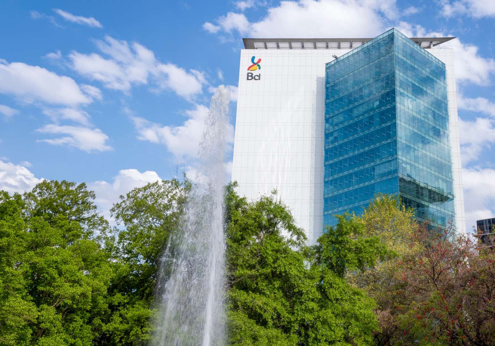Bci's corporate building in glass and white panels, seen from below behind a water fountain and treetops.