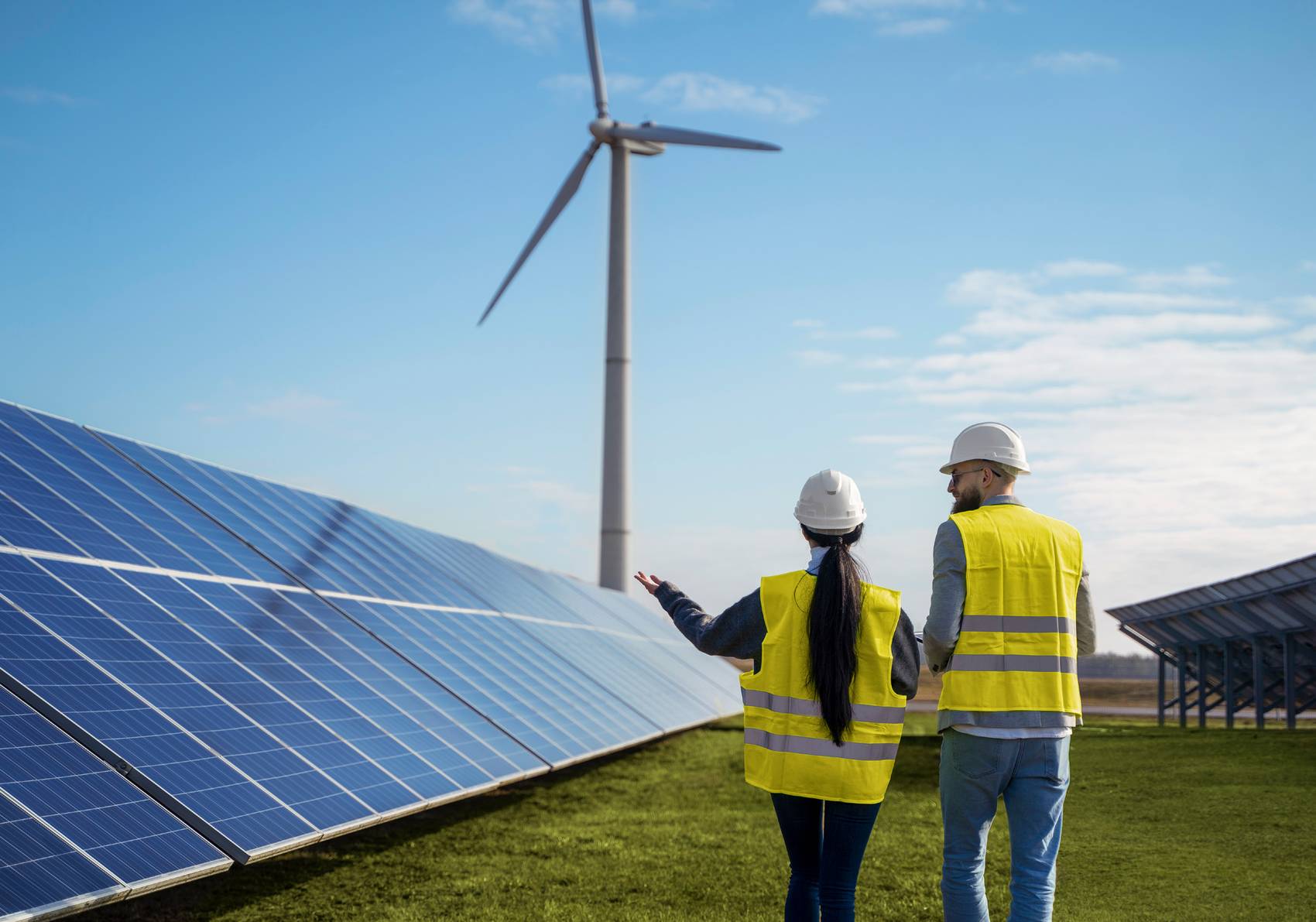 Two technicians in yellow high-visibility vests and hard hats looking at a wind turbine beside rows of solar panels.