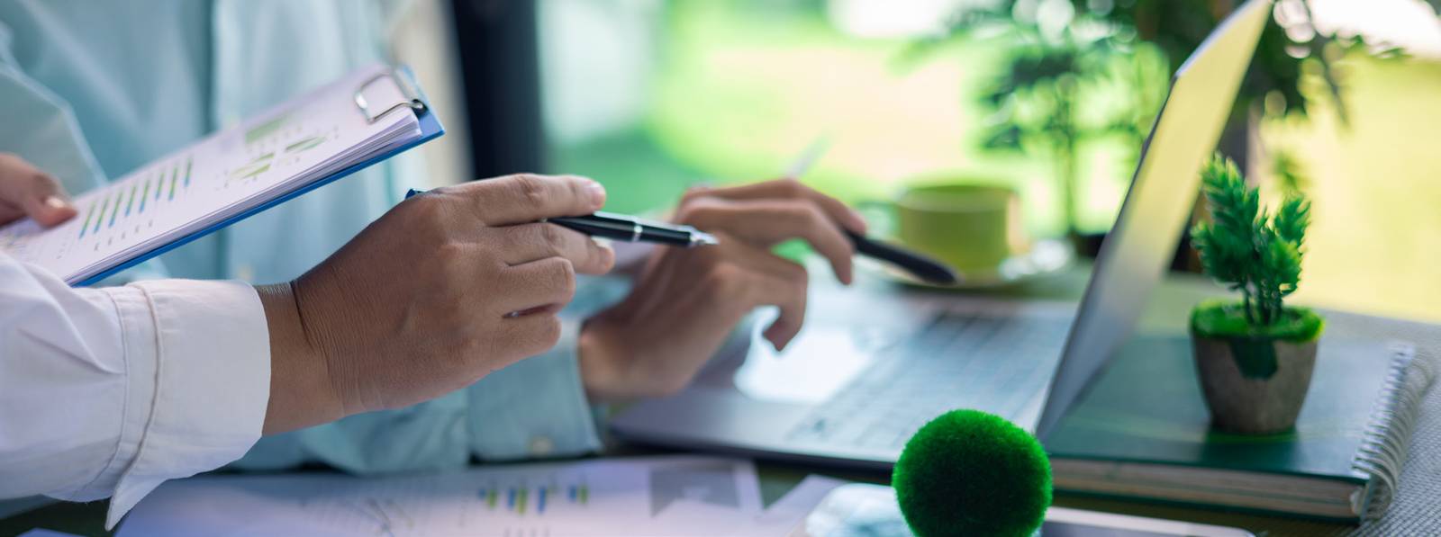 Two people reviewing a printed report with bar charts next to an open laptop and a small potted plant.