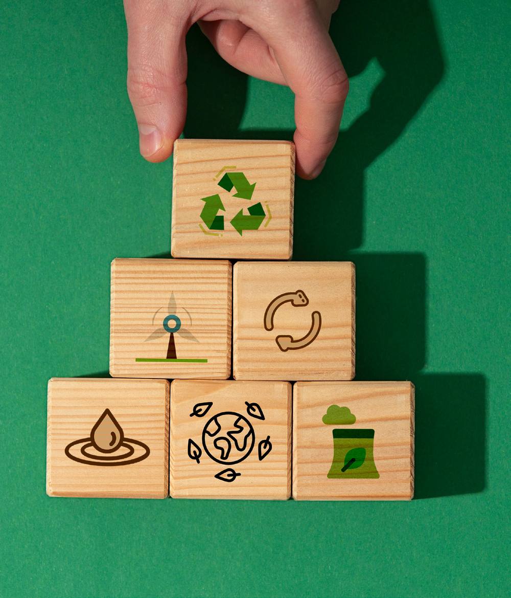 A hand stacking wooden cubes printed with sustainability icons — recycling, wind, water, circularity and clean industry — on a deep green background.